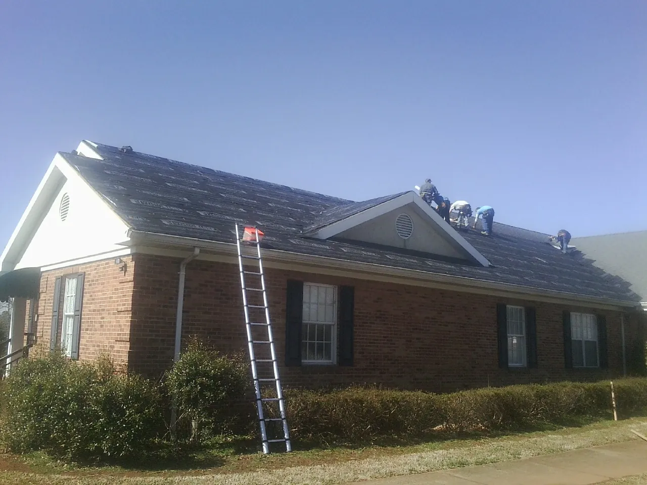 Crew tearing shingles off a brick ranch roof with a ladder set — Emergency Roof Repair work by our crew in Anna Maria