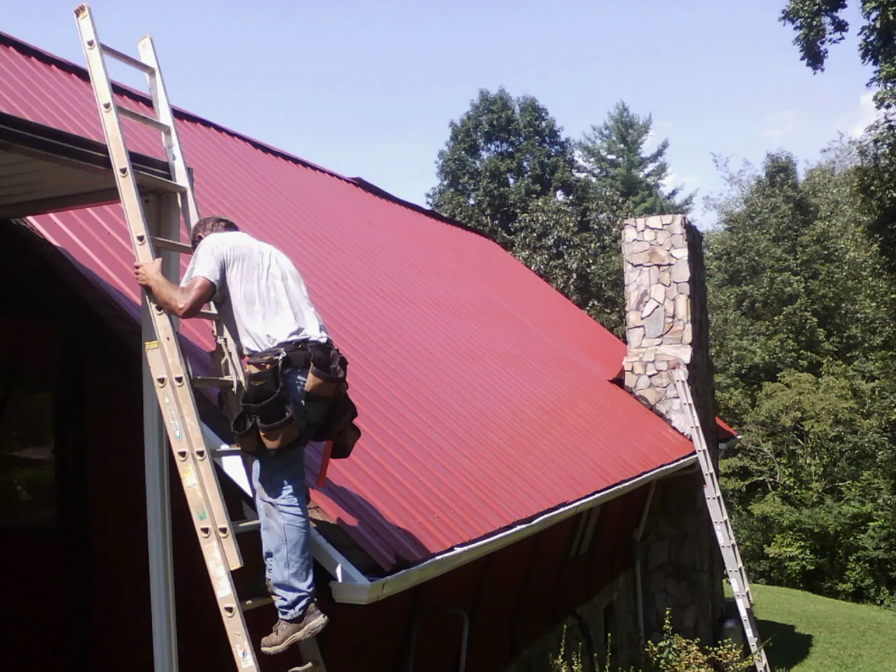 Roofer on a ladder beside a red metal roof and stone chimney — Soffit & Fascia Repair by Roofing Brothers in Anna Maria